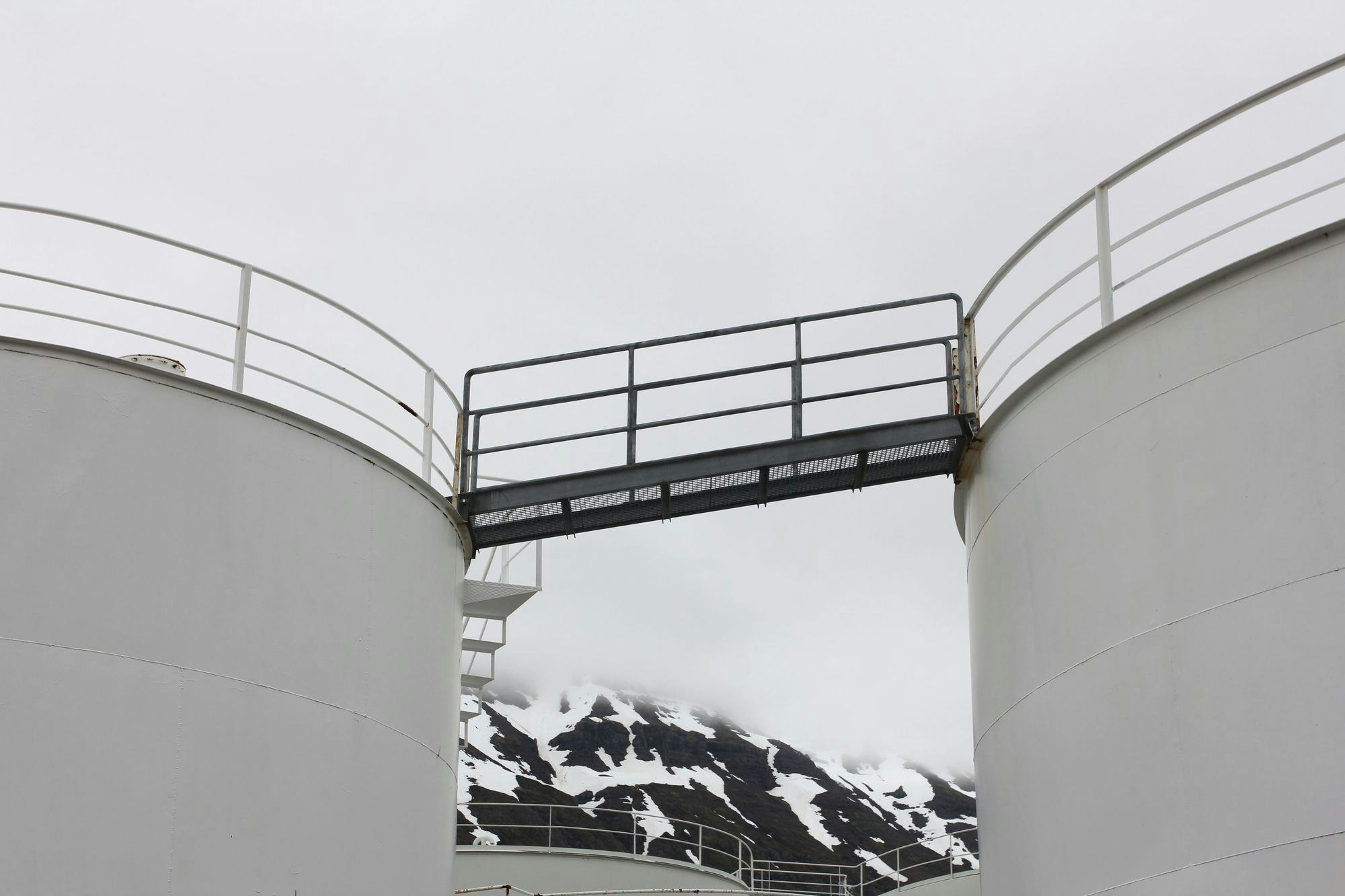 Two white tanks on each side of the photo, a bridge connecting the tanks. Snowy mountains in the back covered with clouds