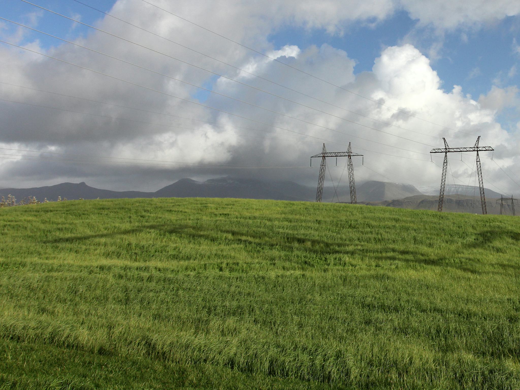 Electricity lines and masts on a green hill, mountains and dramatic clouds in the background
