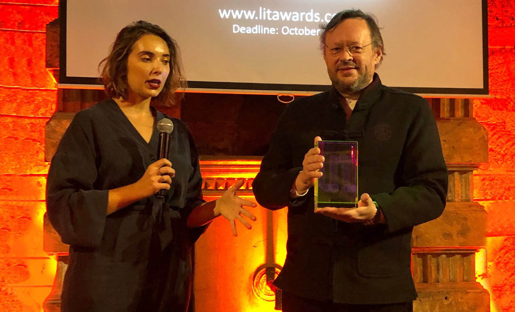 A man holding an award next to a lady with a mic 