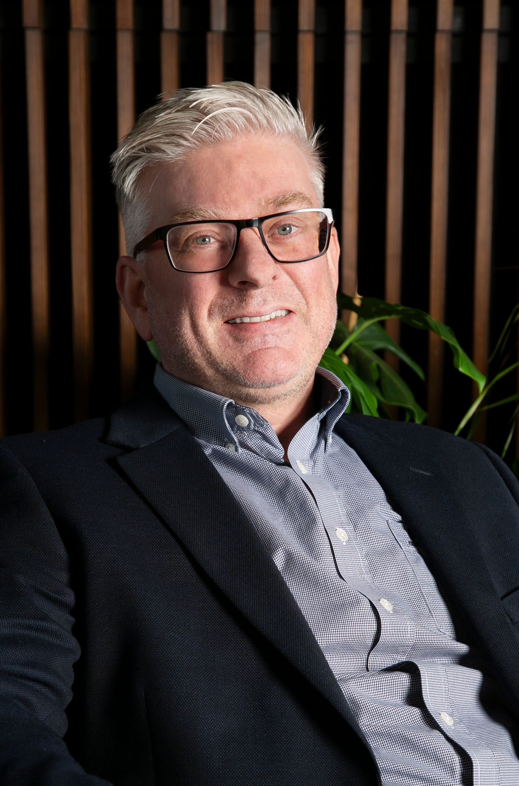 Portrait of a man with glasses smiling, wooden panel and plants in the background