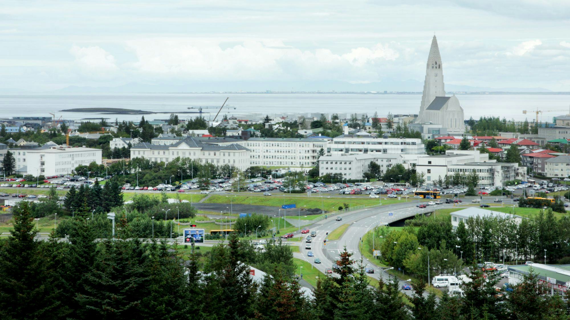 Looking over the city of Reykjavík to the southwest, Hallgrímskirkja church stands out, trees in the foreground, sea in the background
