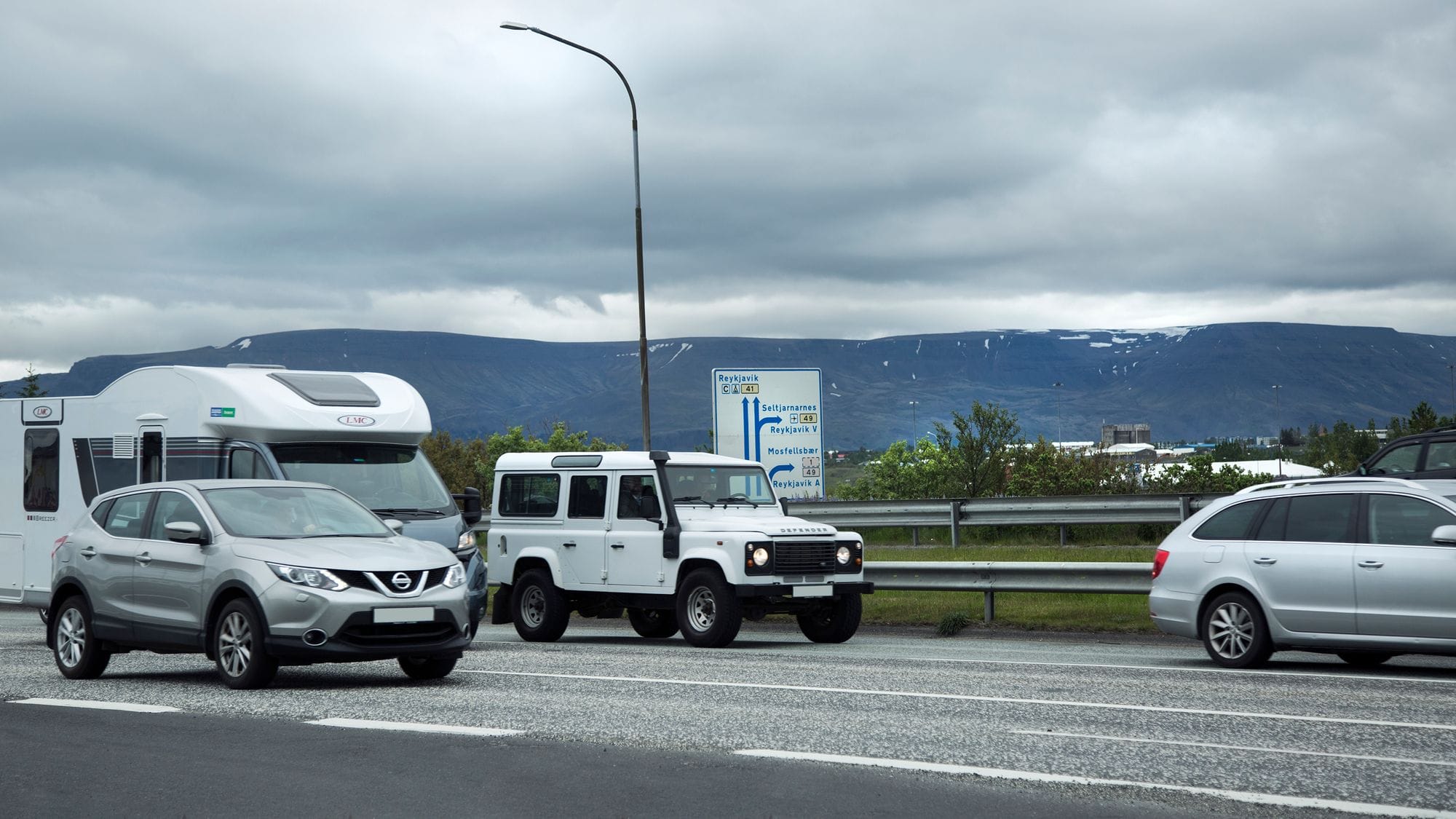 Traffic street, cars drive in two lanes, traffic sign in the middle of the picture, vegetation and houses in the background and at the very back a blue mountain