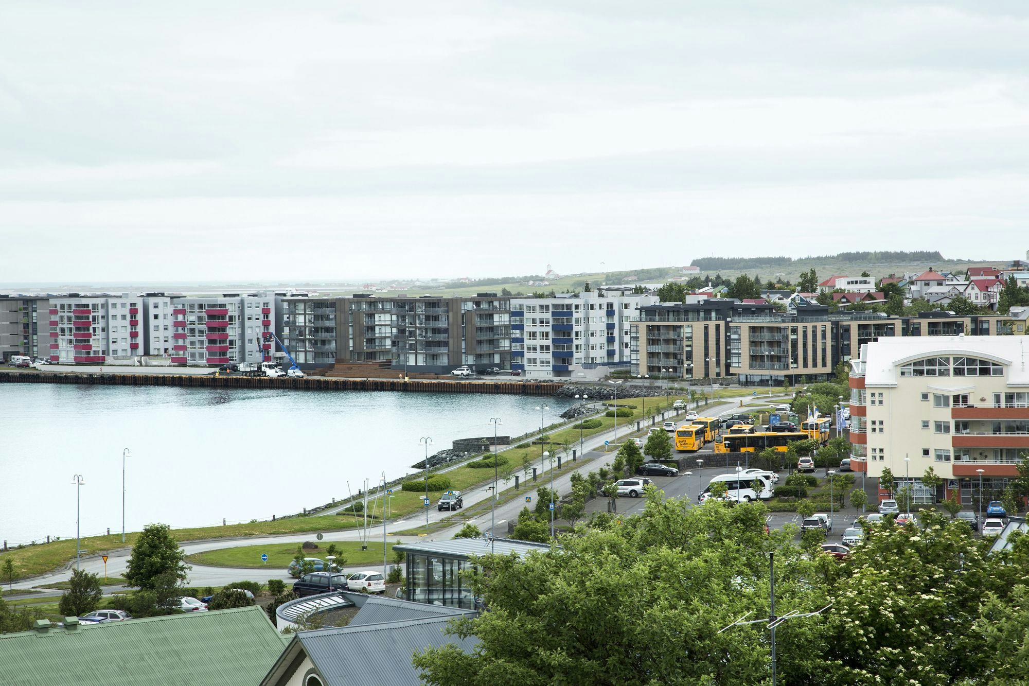 Overview of Hafnarfjörður, apartment blocks by the coastline, the sea and coastline on the left in the foreground, cars and a street running along it, trees and roofs in the foreground