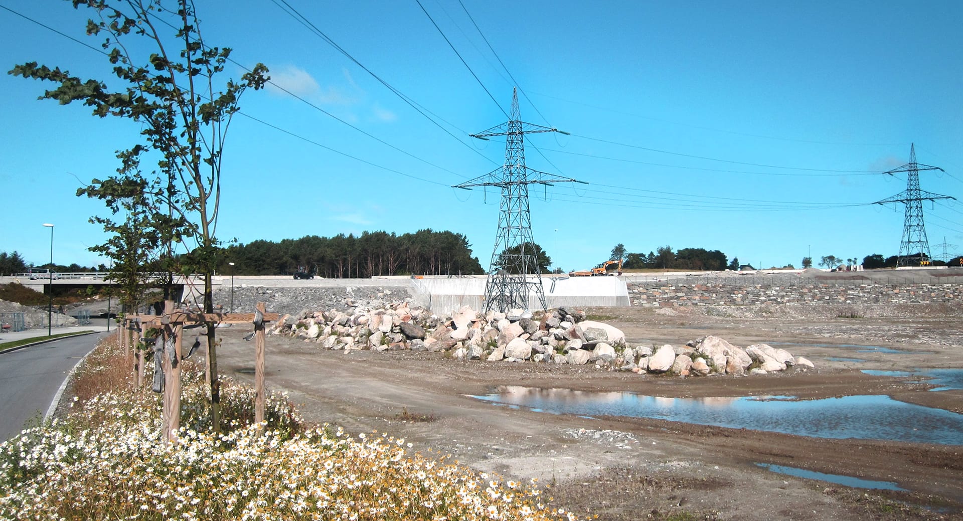 power lines and pylons in suburbia over some kind of riverbed, road on the left and forest in the distance