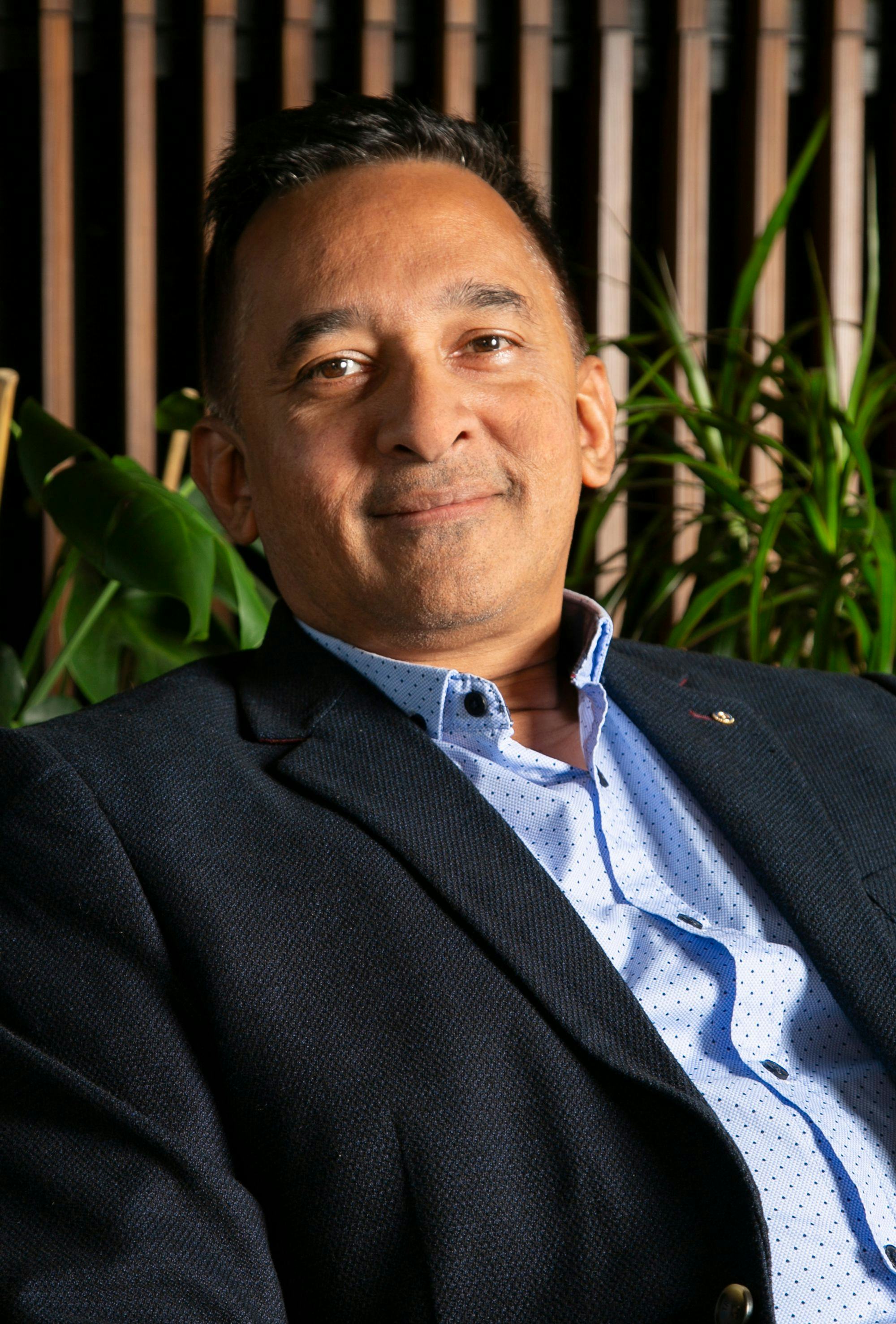 A portrait of a man smiling, wooden panels and plants in the background
